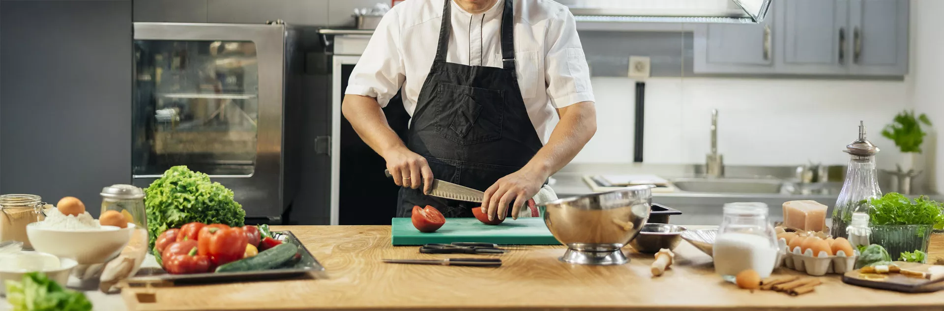 Chef fatiando um tomate em cozinha profissional com ingredientes frescos na bancada, como alface, ovos, farinha, ervas e utensílios de preparo — representando a organização e controle de produção em um restaurante com o CNM.
